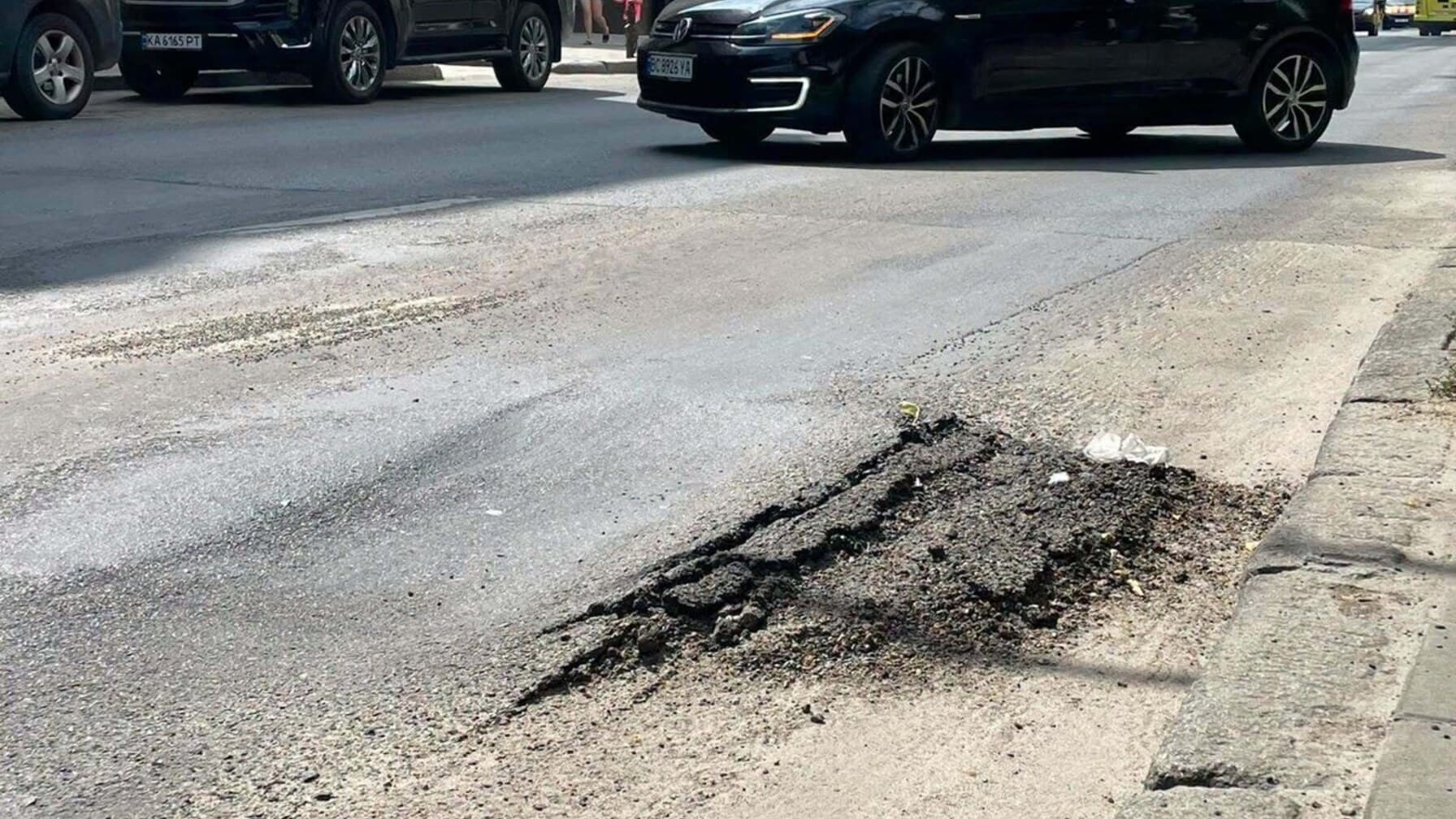 Вулиця Зелена. На передньому плані видно вибоїну та зруйноване дорожнє покриття — асфальт потріскався, частина поверхні просіла. На задньому плані рухаються автомобілі, поруч розташовані будинки та тротуар. / Фото: ЛМР